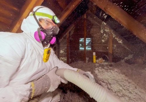 A weatherization technician in protective suit, respirator, and goggles installs insulation with a hose in an attic space.