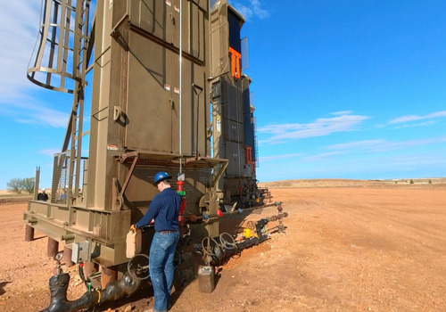 A worker in a hard hat stands inspects an oil pumping unit in a vast field.