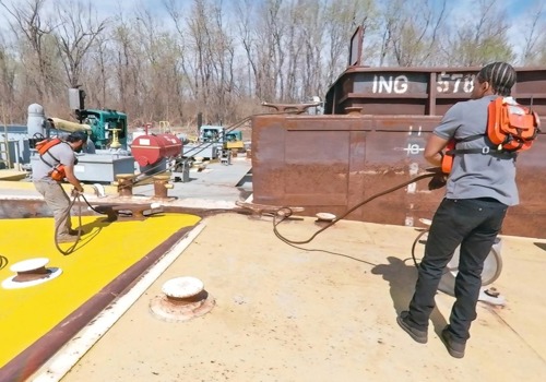 Two deckhands wearing safety vests work on a barge deck, using rope to tether the vessel to another on a sunny day.