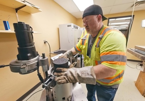 Engineering technician in safety gear tests a cylindrical asphalt sample using lab equipment.